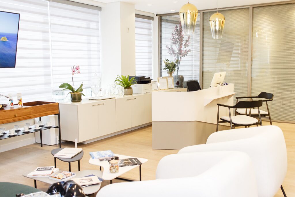 Recepção da Clínica Rafaela Salvato Dermatologia com balcão branco curvo, poltronas e luminárias douradas em ambiente claro e moderno.